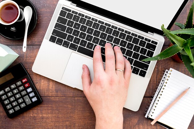 Close-up of a person's hand typing on laptop with calculator; coffee cup and spiral notepad with pencil on wooden desk