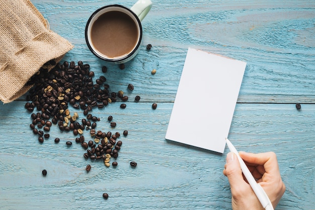 Close-up of a person's hand writing on blank paper with cup of a coffee and coffee beans