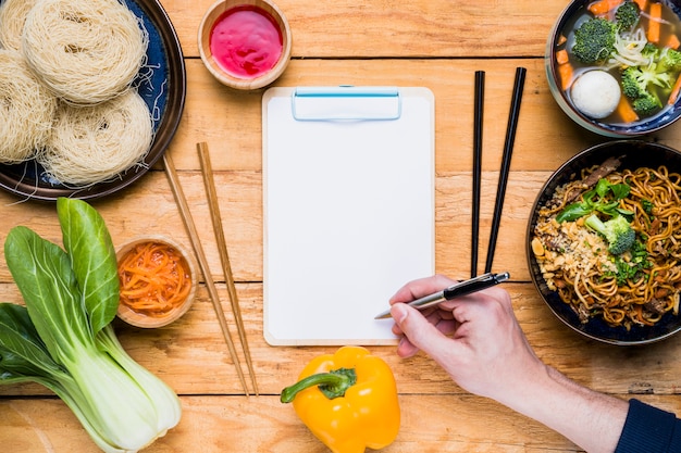 Close-up of a person's hand writing on clipboard with traditional thai food on wooden table