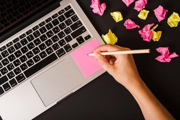 Close-up of a person's hand writing on pink adhesive note on laptop against black background