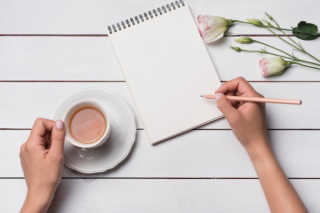 Primo piano di una persona che scrive sul blocco note con la tazza di tettarella sulla scrivania in legno