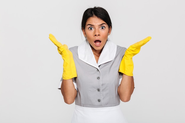 Close-up photo of amazed young female housekeeper in uniform and protective gloves standing with opened palms