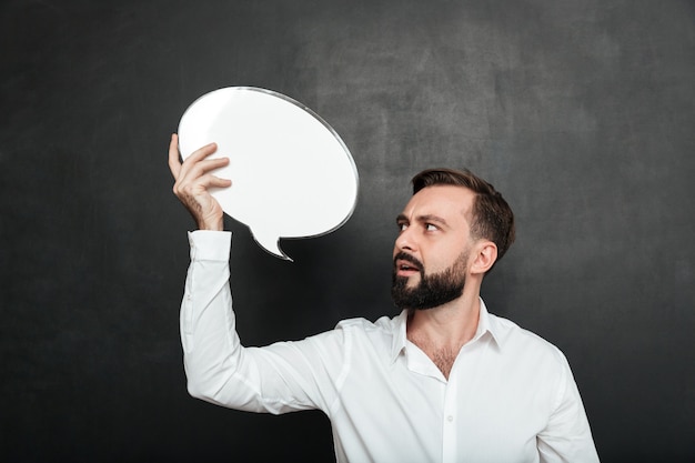 Close up photo of attractive man holding blank message ballon and looking at announcement over dark gray wall copy space