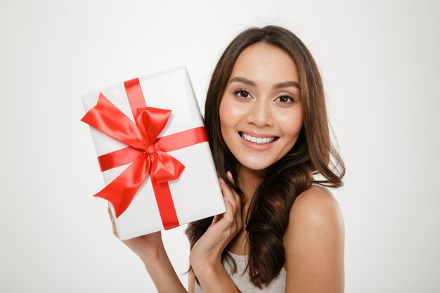Close up photo of cheerful woman showing gift-wrapped box with red bow on camera expressing happiness and delight, isolated over white