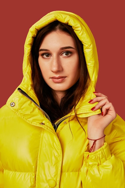 Free photo close up photo of cute girl with dark hair in studio, in bright yellow down jacket. red background, oversized clothes, hood on