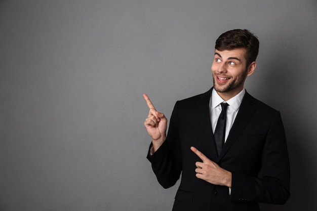 Close-up photo of handsome bearded man in black suit poiting with rwo fingers, looking aside