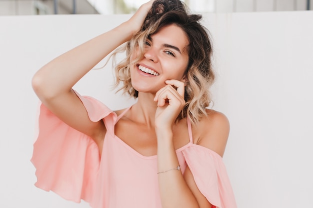 Close-up photo of inspired tanned girl playing with blonde wavy hair. Portrait of dreamy caucasian female model wears trendy pink clothes.