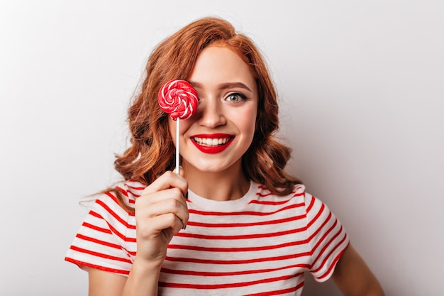 Close-up photo of jocund ginger woman with red candy. Joyful european girl with curly hair eating lollipop.