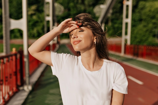 Close up photo of lovable cute lady with dark wavy hair wearing white tshirt is posing with lovely smile and closed eyes on bright bridge in sunlight