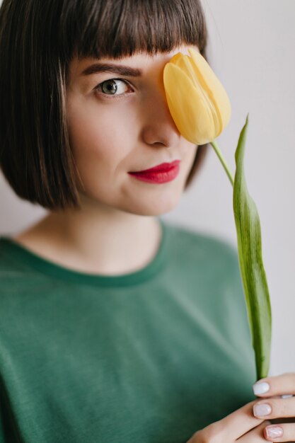 꽃과 함께 포즈를 취하는 갈색 머리를 가진 사랑스러운 유럽 여자의 클로즈업 사진. 노란 튤립과 기쁘게 세련 된 여자의 실내 초상화.