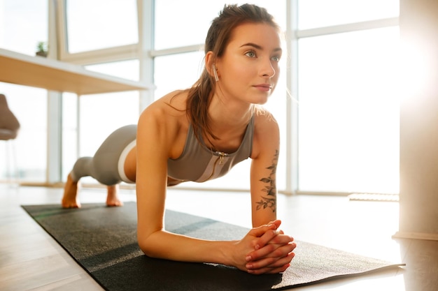 Close up photo of pretty lady in sporty top and leggings practicing fitnes and listening music in earphones at home with big windows on background.