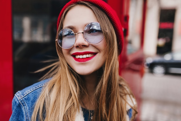 Крупным планом фото романтичной белой девушки носит круглые очки, глядя с улыбкой. Мечтательная барышня с ярким макияжем позирует возле красного автобуса.