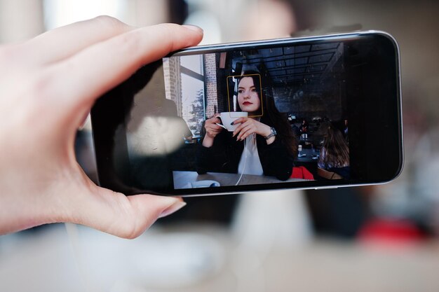 Close up photo of screen mobile phones with brunette girl sitting on cafe with cup of cappuccino listening music on headphones