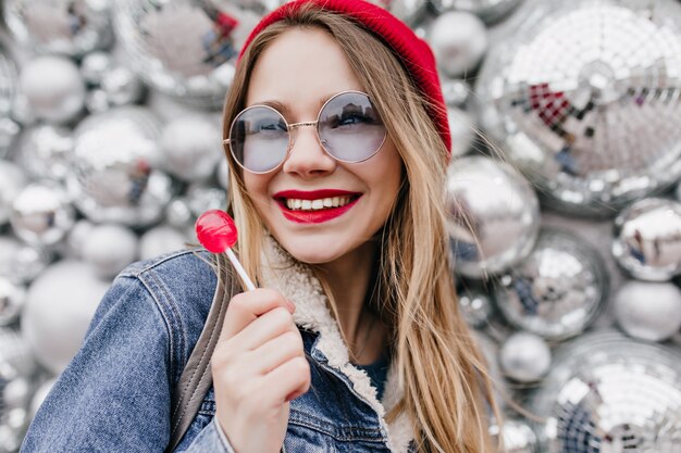 Close-up photo of smiling girl in denim jacket posing with red lollipop. Portrait of amazing white female model standing near disco balls with candy.