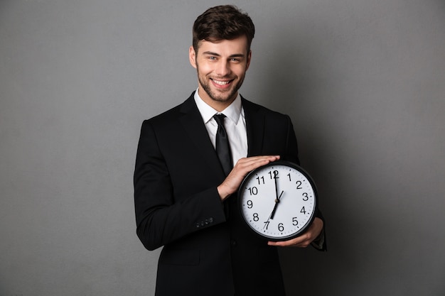 Close-up photo of smiling handsome man in black suit holding clock, 