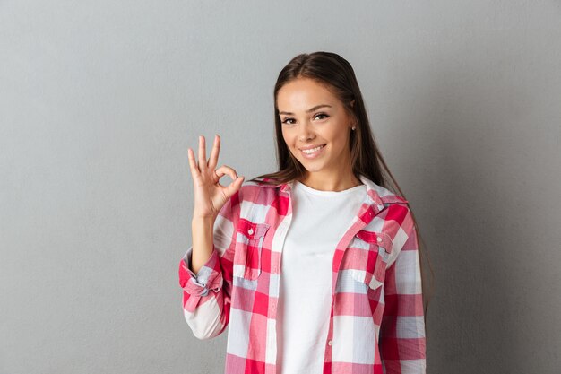 [ok]ジェスチャーを示す市松模様のシャツで笑顔の若いブルネットの女性の写真をクローズアップ