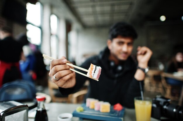 Крупный план фото палочек, держащих случайного и стильного молодого азиата с наушниками в кафе, поедающем суши