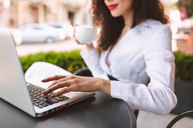 Close up photo of woman body sitting at the table with cup of coffee in hand and laptop in cafe on street