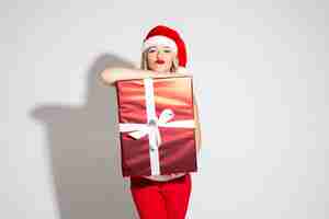 Free photo close-up photo of a young blonde lady wearing santa hat and red lipstic while leaning on a christmas present. holiday concept