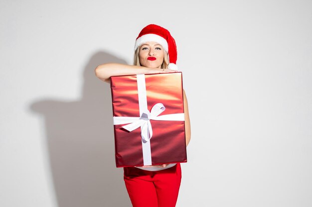 Крупным планом фото молодой блондинки в шляпе Санты и красной помаде, опираясь на рождественский подарок. Концепция праздника