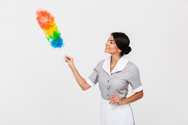 Close-up photo of young cheerful housekeeper in uniform cleaning with duster, looking aside