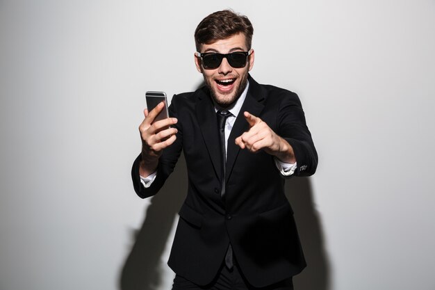 Close-up photo of young happy exited attractive man in sunglasses holding smartphone and pointing with finger on you