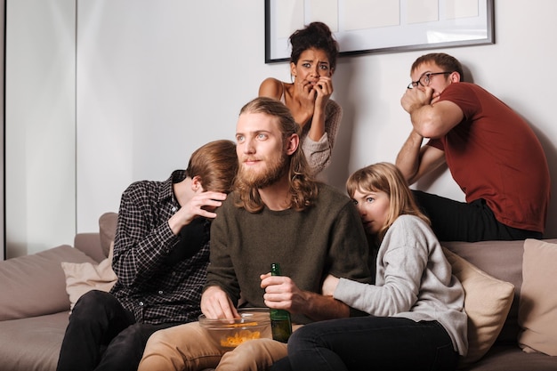 Close up photo of young smiling man and his scared friends sitting on sofa with chips and beer and watching horror movie together at home