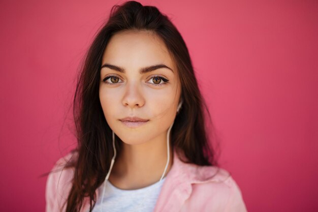 Close up photo of young thoughtful girl with dark hair standing and listening music in earphones while dreamily looking in camera on pink background