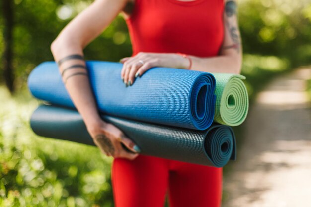 Close up photo of young woman body in red jumpsuit standing with colorful yoga mats in hands in park