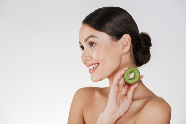 Close up picture of smiling woman with healthy fresh skin holding kiwi and looking aside, isolated over white