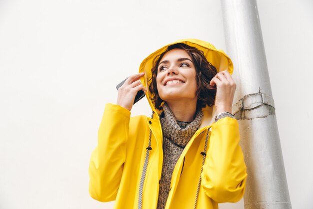 야외에서 걷고있는 동안 후드를 착용하고 비오는 날씨를 즐기는 노란 코트에 세련된 여성의 사진을 닫습니다