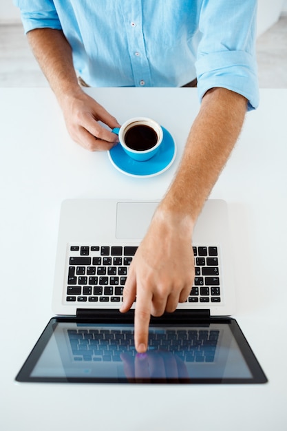 Close up picture of young businessman's hands sitting at table pointing on laptop screen holding coffee cup. White modern office interior 