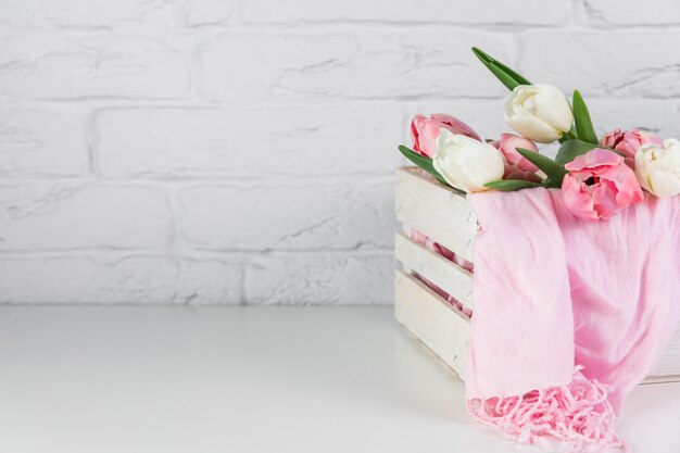 Close-up of pink scarf and tulips flower inside the wooden crate on desk against brick wall