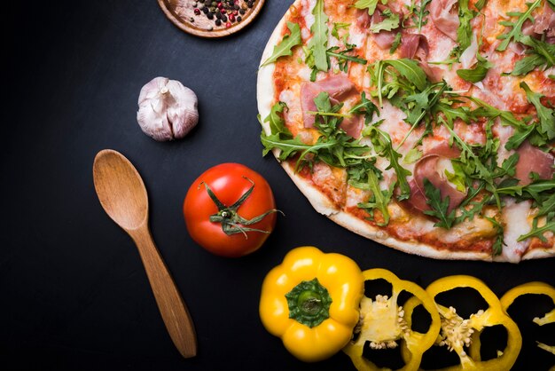 Close-up of pizza with arugula leaves; slices of yellow bell pepper; tomato and garlic bulb over black background