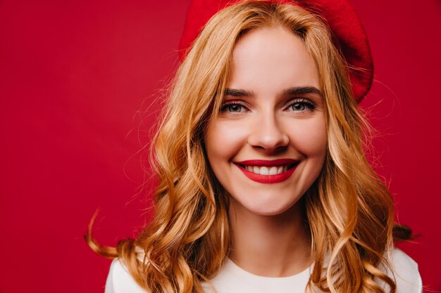 Close-up portrait of adorable blonde woman isolated on red wall. Laughing amazing girl posing in french beret.