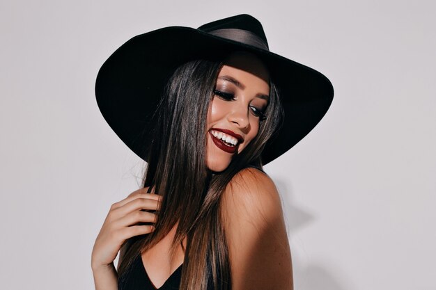 Close up portrait of adorable gothic young woman in witch halloween costume with hat standing and smiling over white isolated wall