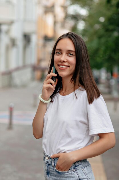 하얀 티셔츠와 청바지를 입고 행복한 미소와 검은 머리를 한 사랑스러운 사랑스러운 여성의 초상화를 닫고 흐릿한 도시 배경에서 전화 통화를 하고 웃고 있습니다