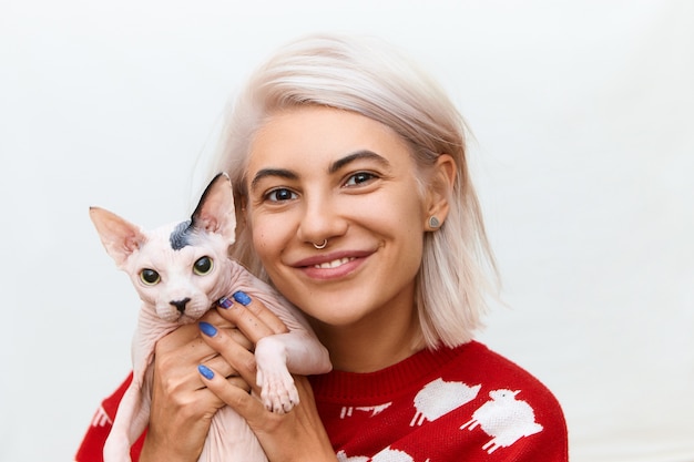 Close up portrait of adorable pretty girl with facial piercing posing with her four legged best friend. Cheerful happy young female with dyed hair embracing Sphynx cat with no fur coat