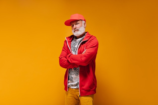 Close up portrait of adult man in red outfit on orange background cool handsome guy with grey beard in cap and bright sweatshirt posing