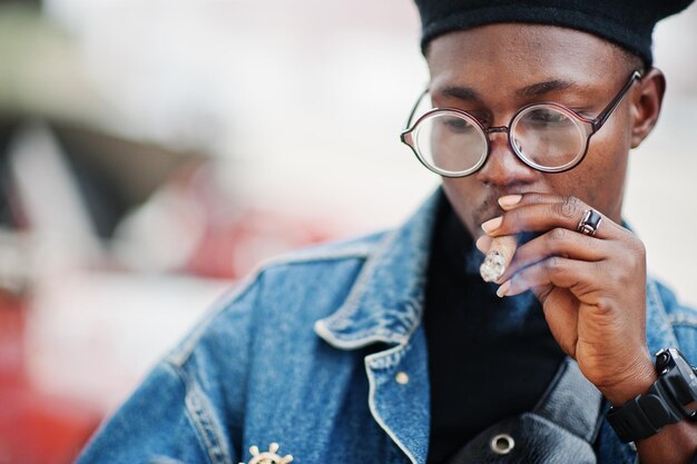 ジーンズジャケットベレー帽と眼鏡喫煙葉巻でアフリカ系アメリカ人の男の肖像画をクローズアップ