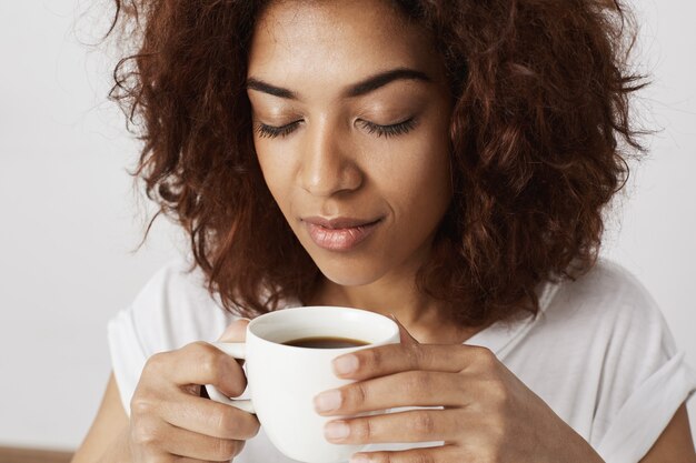 目を閉じてコーヒーの香りのカップを保持しているアフリカの女の子の肖像画を閉じます。朝目を覚ますことは大人のために厳しいです。