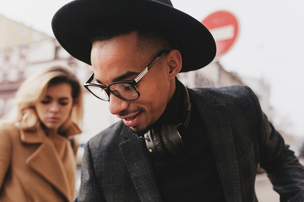 Close-up portrait of african man looking down while posing with blonde woman. Joyful black male model in hat spending leisure time with euopean girl.