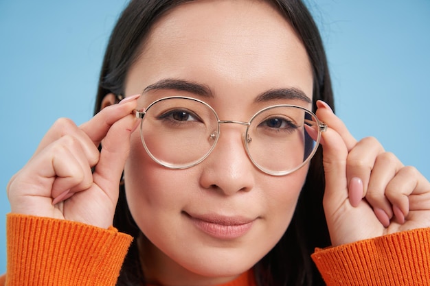Close up portrait of amazed girl looks closer at camera in glasses standing against blue background
