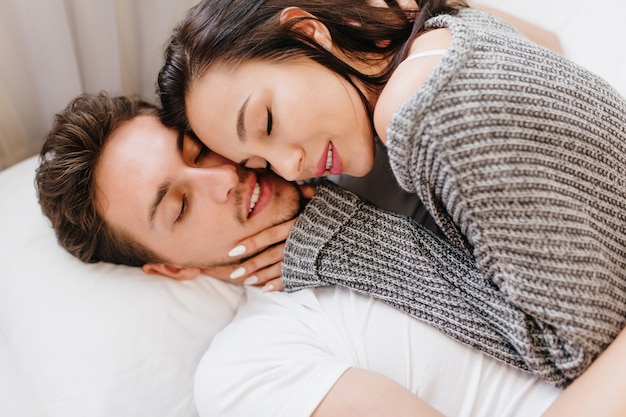 Close-up portrait of amazing brunette female model lying in bed with husband in white t-shirt