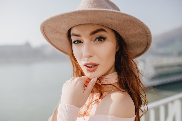 Close-up portrait of amazing ginger woman in beige fedora standing on sea