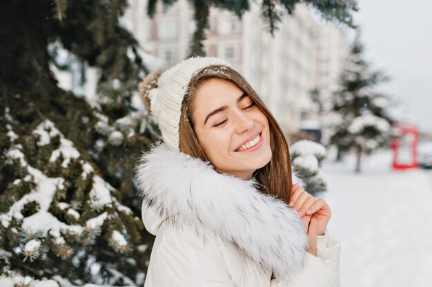 都市の冬時間を楽しんでいる白い暖かい暖かい服で素晴らしいうれしそうな女性の肖像画を間近します。目を閉じて笑っている雪の中で若いきれいな女性。