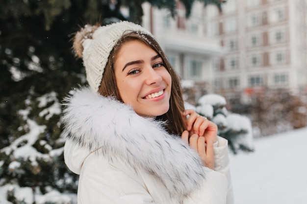 Close-up portrait of amazing woman in white knitted hat with snowflakes on hair smiling on street.