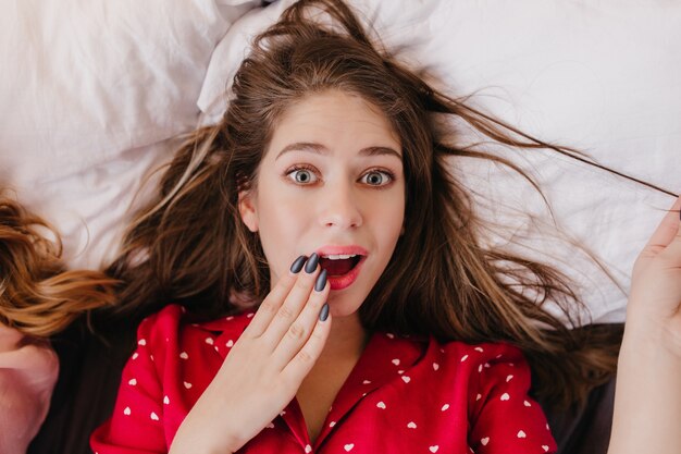 Close-up portrait of amazing young woman wears stylish red pajamas. Indoor overhead shot of surprised brunette girl lying on white pillow.