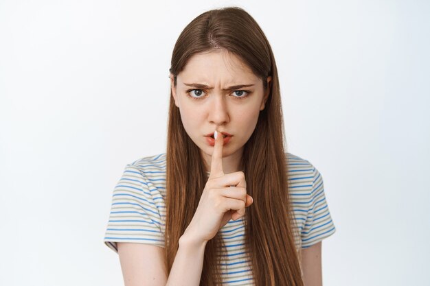 Close up portrait of angry girl shushing, hush gesture, frowning and press finger to lips, warn someone, telling to be quiet on white