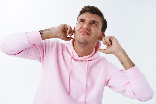 Free photo close-up portrait of annoyed and disturbed blond man in pink hoodie cant work from home during quarantine covid19 cause neighbour upstairs being too loud, shut ears not hear noisy music